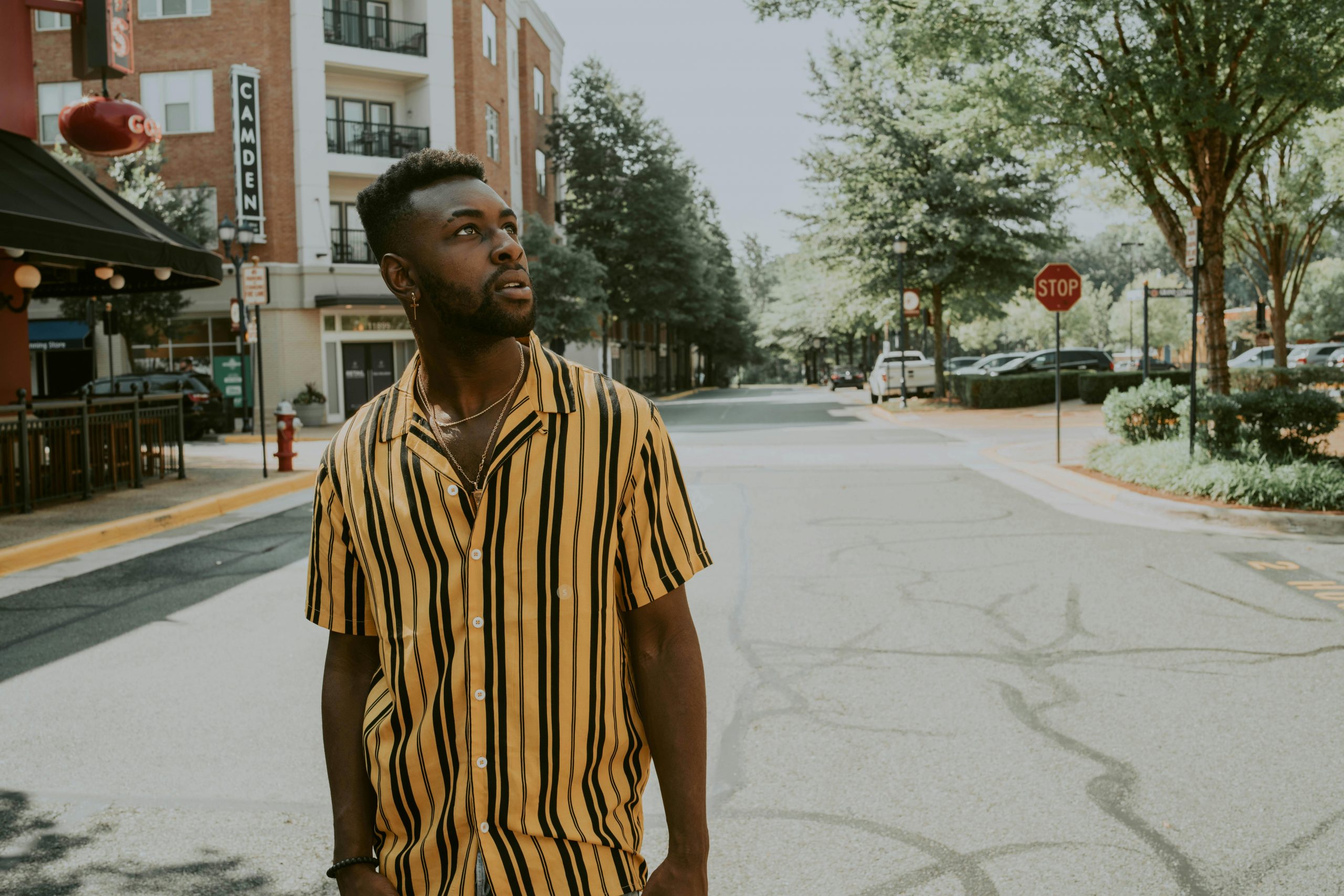 Young stylish dreamy African American guy in bright striped shirt and creative haircut looking up on asphalt roadway